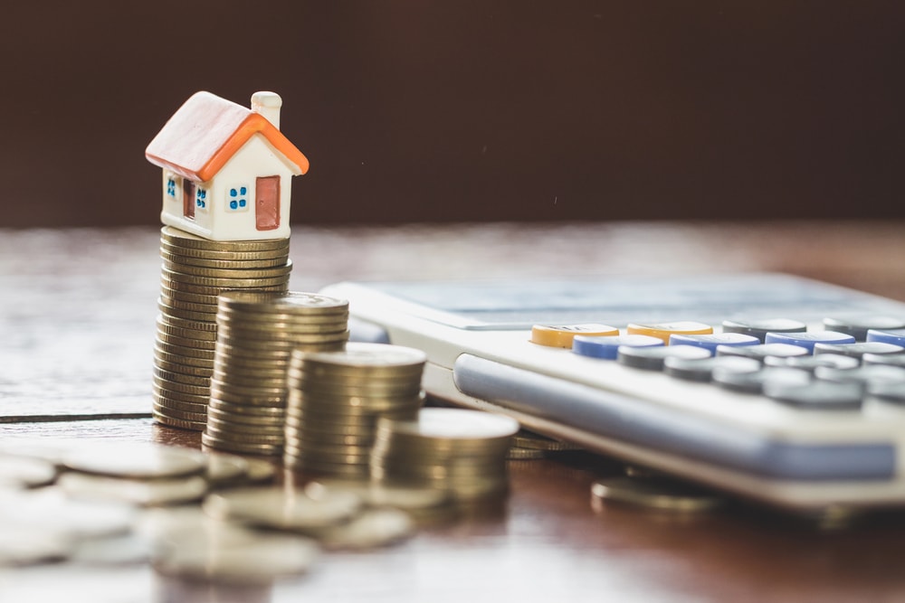 a mock house sitting on a stack of coins by a calculator, illustrating property tax laws and burden